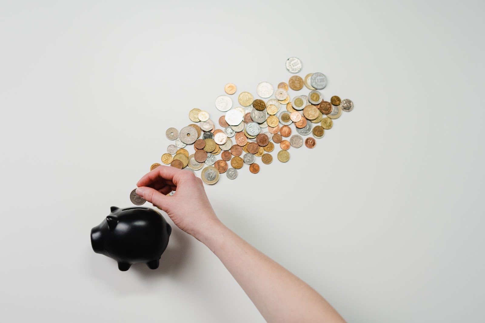 Hand dropping a coin into a black piggy bank beside a trail of coins